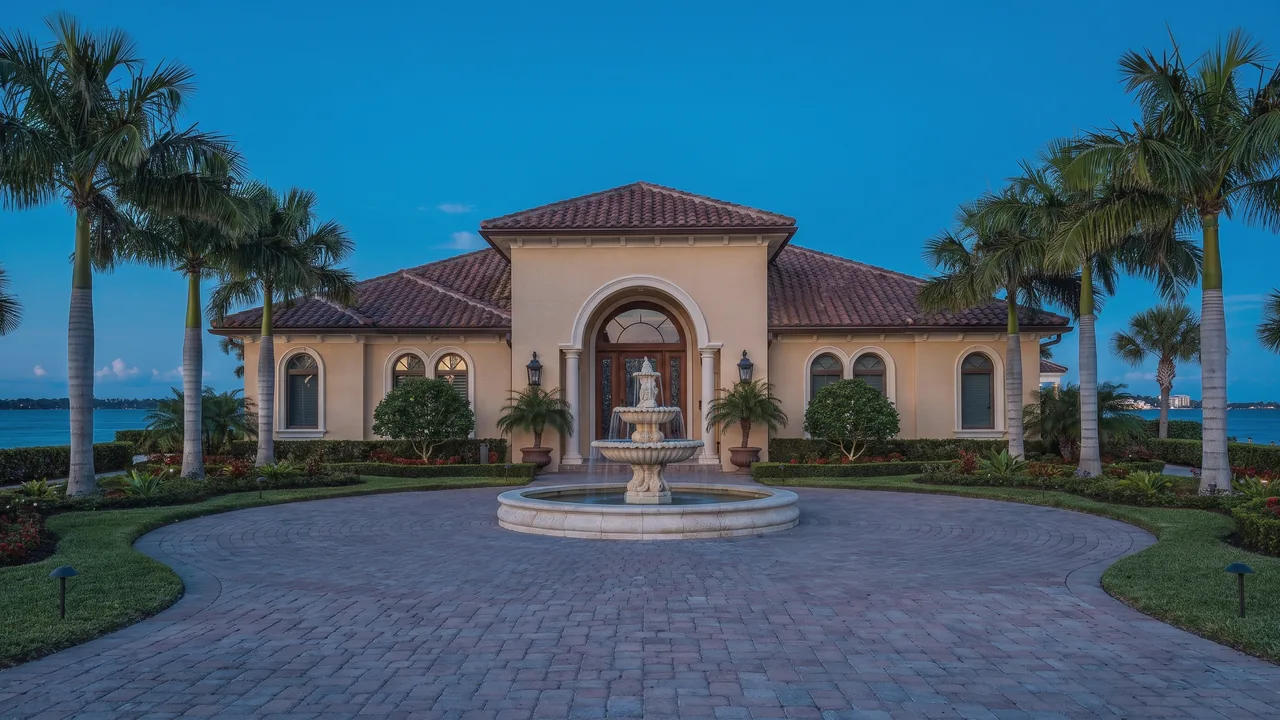 A waterfront house at dusk with a fountain in a paved motor court, framed by palms