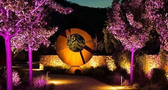 Trees lit in magenta beside a sculpture lit in warm amber in a garden at night