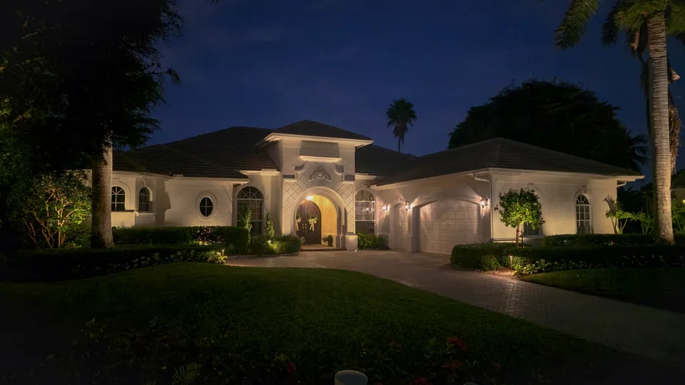 A white home at dusk with its arched entry, facade, planting and drive lit