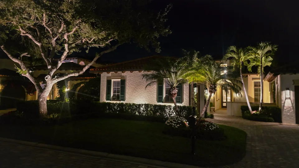 A Mediterranean-style house with an uplit oak, lit palms and a washed elevation behind a lawn