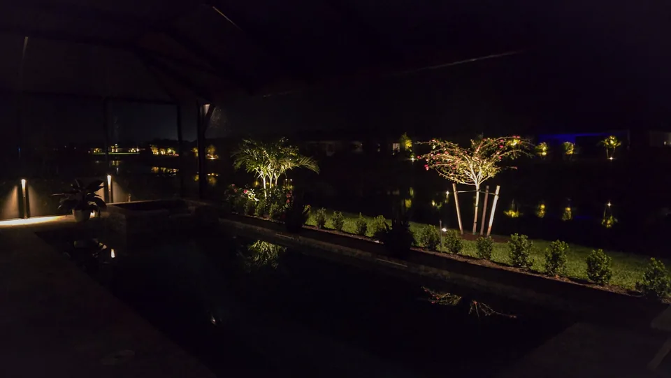 Bollard lights inside a lanai and a lit planting bed of palms and bougainvillea beyond the pool