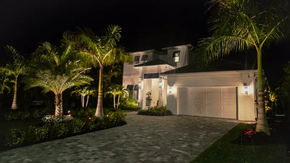 A modern two-story home at night with palms uplit along a paver drive and lanterns at the entry