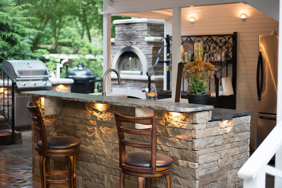 An outdoor kitchen with a stone bar, grill and pizza oven, lit under its counter ledge