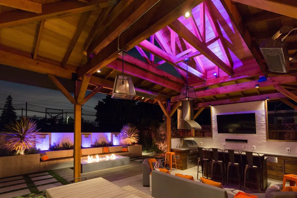 A covered outdoor room at night with a kitchen, a bar and a fire feature