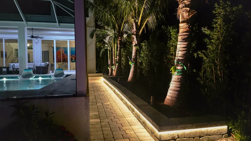 A paver walk lit by a continuous strip tucked under a wall cap beside a pool