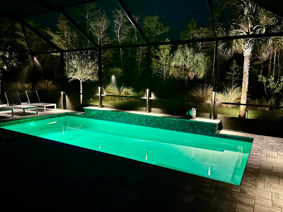 A screened pool enclosure at night with low lights at the deck edge and lit planting outside the screen