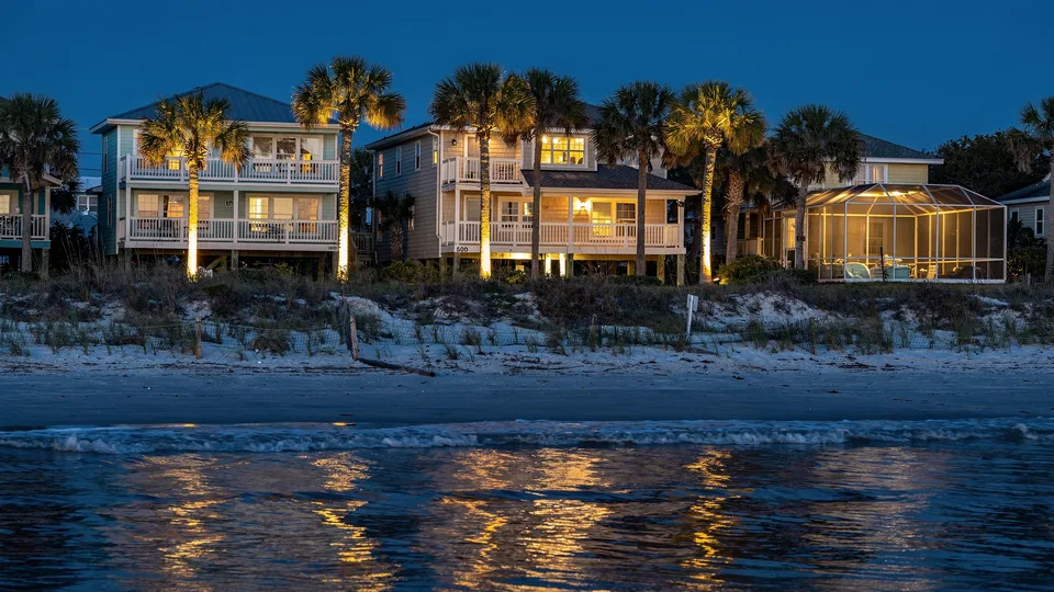 Illustration of beach homes behind a dune at dusk with uplit palms and a lit pool cage