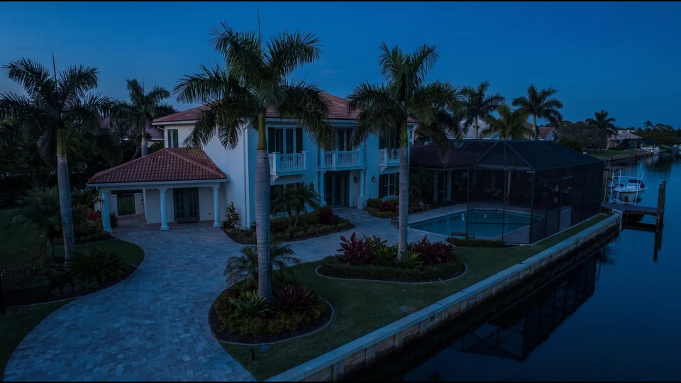 An aerial view of a waterfront house at dusk: drive and entry, palms, planting beds, a screened pool and a lit seawall on a canal